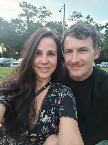 Couple smiling, posing close together outdoors; woman in floral print dress, man in black shirt.