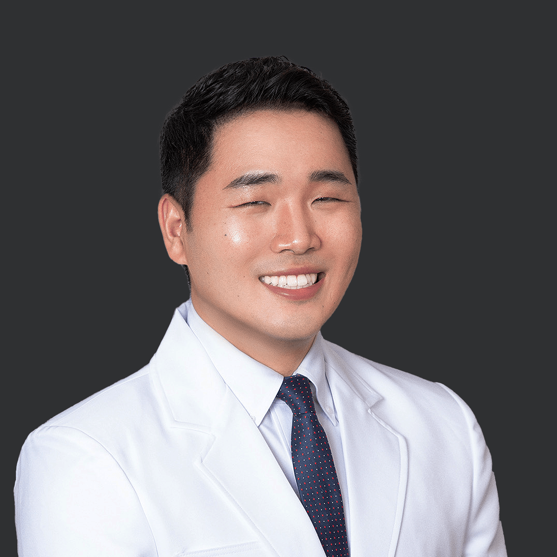 A smiling person wearing a white lab coat and a dark blue tie, set against a dark gray background.
