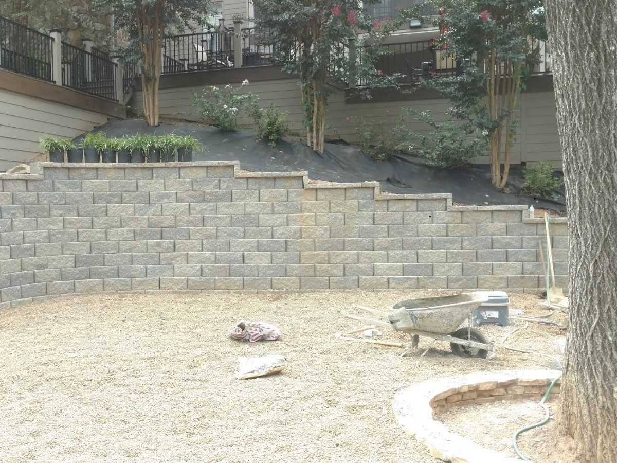 Retaining wall of gray blocks on a slight hill, with a gravel yard and trees.