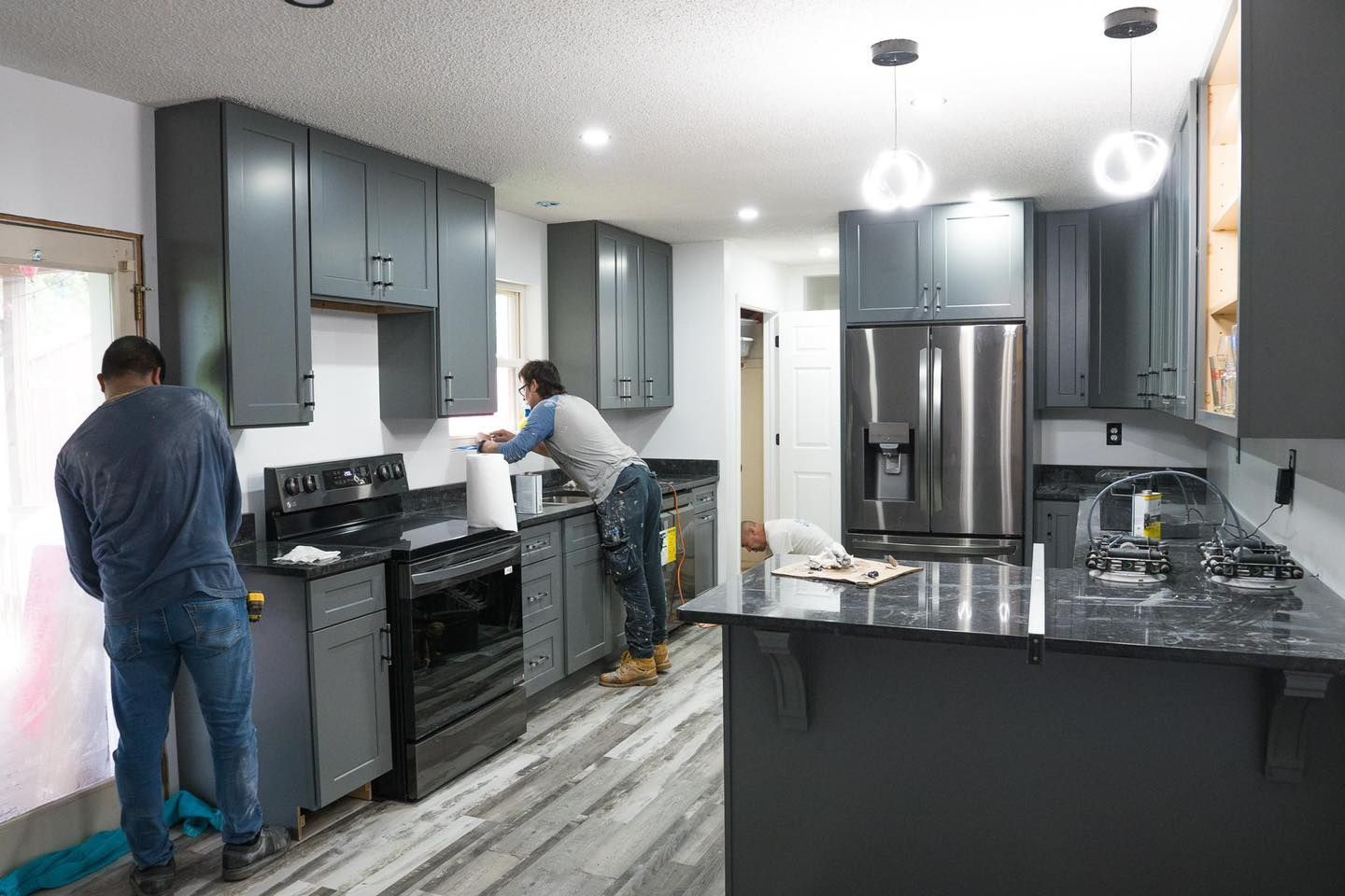 Two men installing cabinets in a remodeled kitchen with gray cabinets, black appliances, and a stainless steel refrigerator.