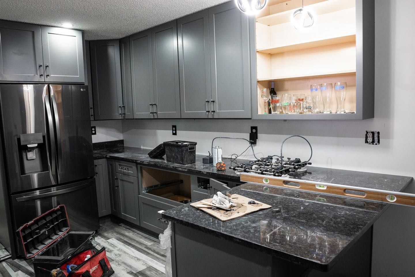 Kitchen renovation: gray cabinets, black countertops, stainless steel fridge, tools, exposed cabinetry, and unfinished walls.