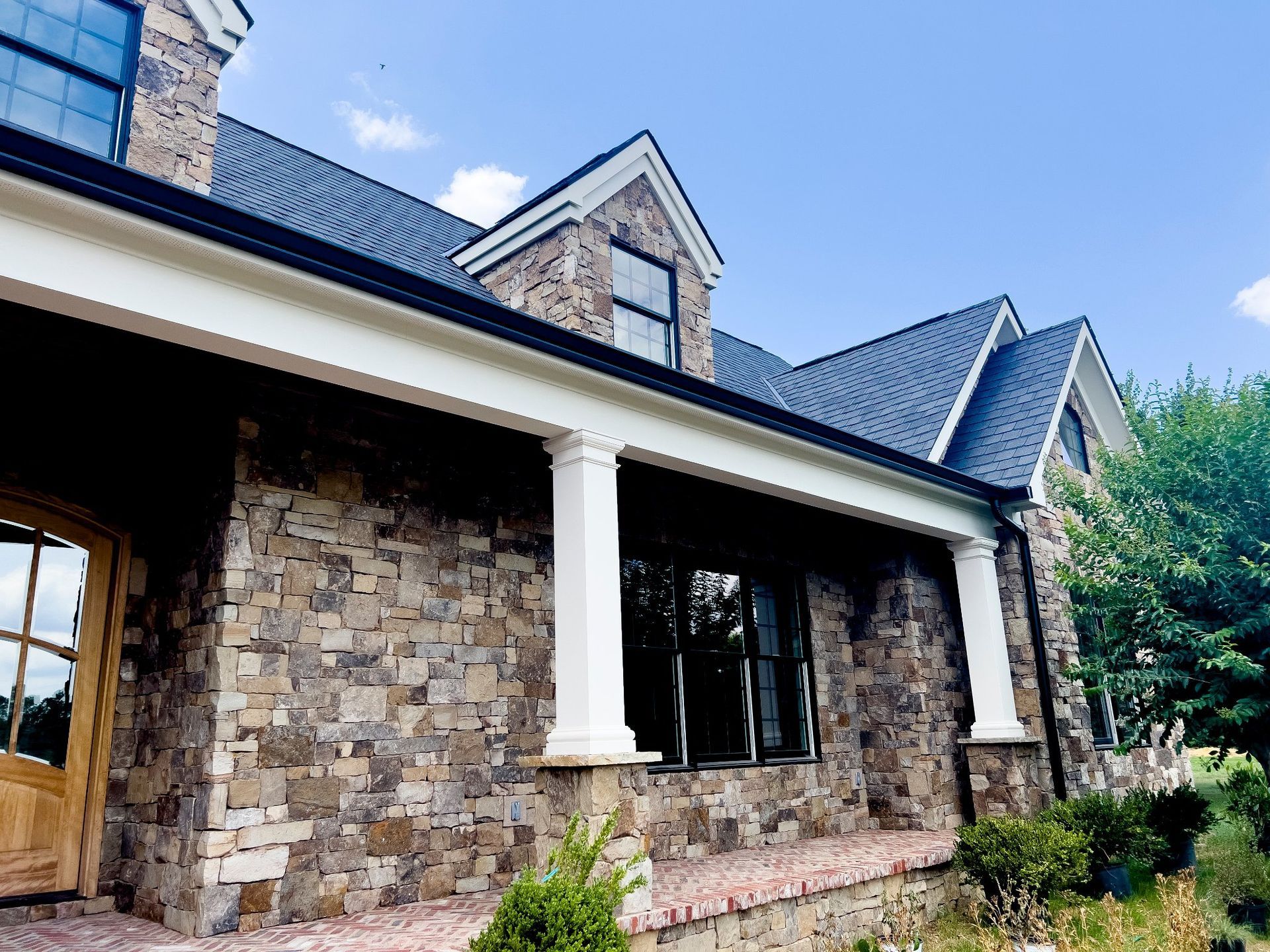 Stone-faced house with white columns, black roof, and windows against a blue sky.