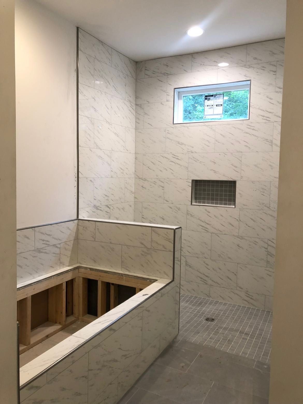 Bathroom with marble-look tiled walls, shower with built-in niche, unfinished tub area, small window, and grey floor.