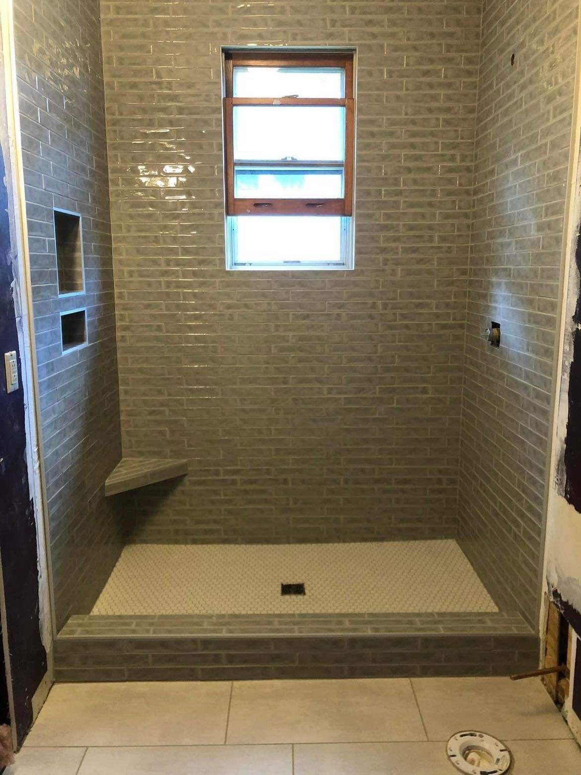 A newly tiled shower stall with grey brick-like tiles, a window, built-in shelf, and a white tiled floor.