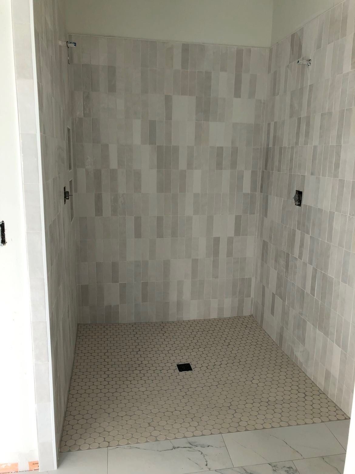 Shower stall with light-colored rectangular tiles on the walls and small, circular tiles on the floor.