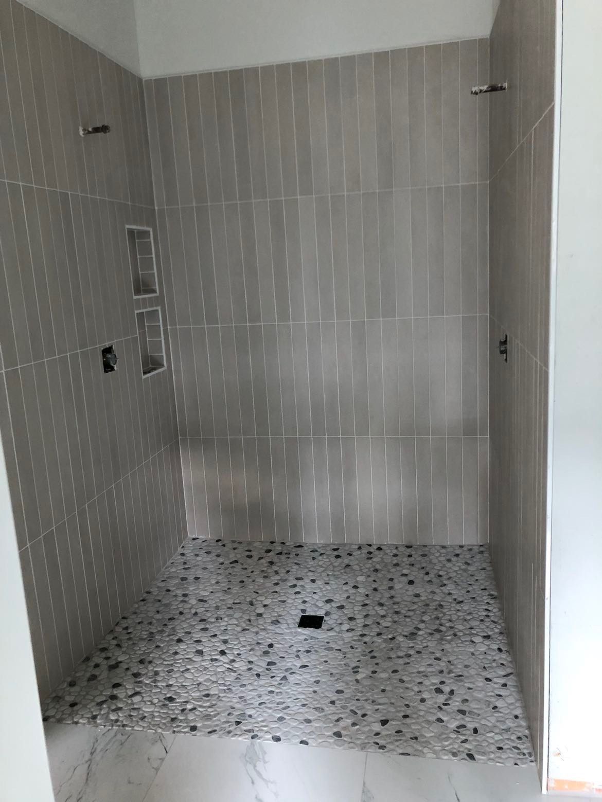 Completed shower with gray tile walls, pebble floor, and recessed shelf.