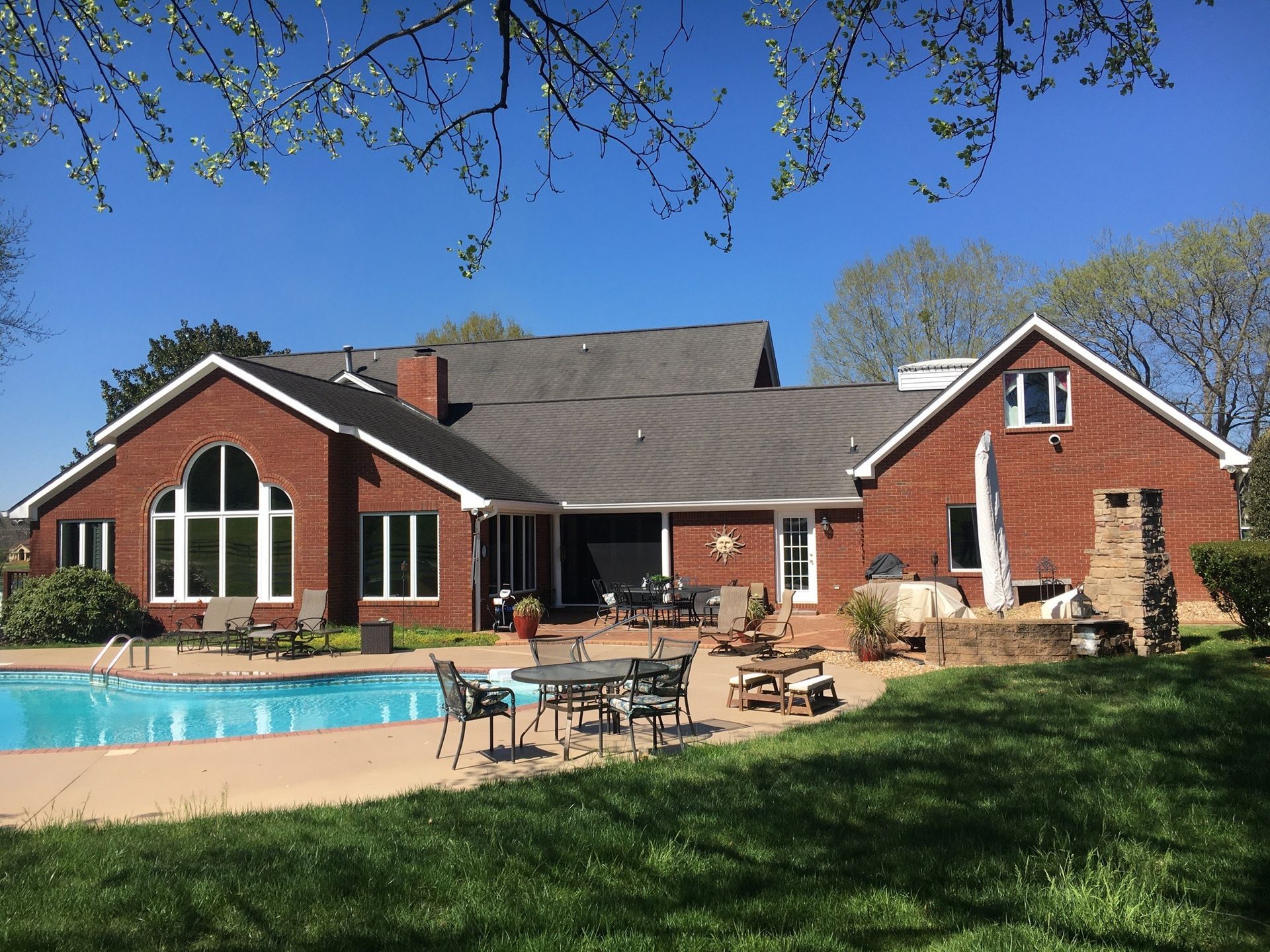 Red brick house with a pool, patio furniture, and a stone fireplace on a sunny day.