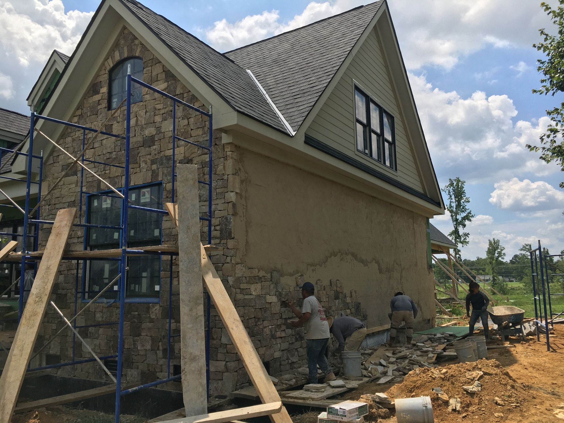 Construction workers applying stone siding to a house. Scaffolding, mortar, and materials are visible.