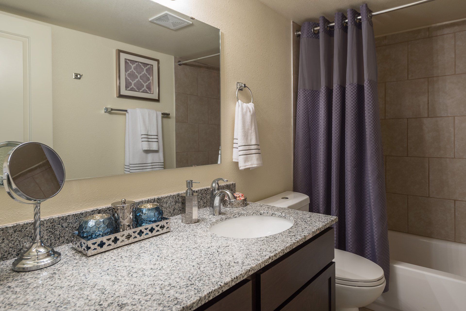 Small bathroom with speckled vanity, mirror, towels, and purple shower curtain beside toilet.