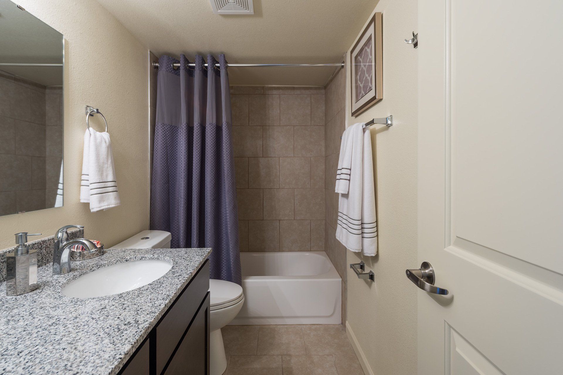 Small bathroom with granite vanity, toilet, tiled tub shower, and gray curtain.
