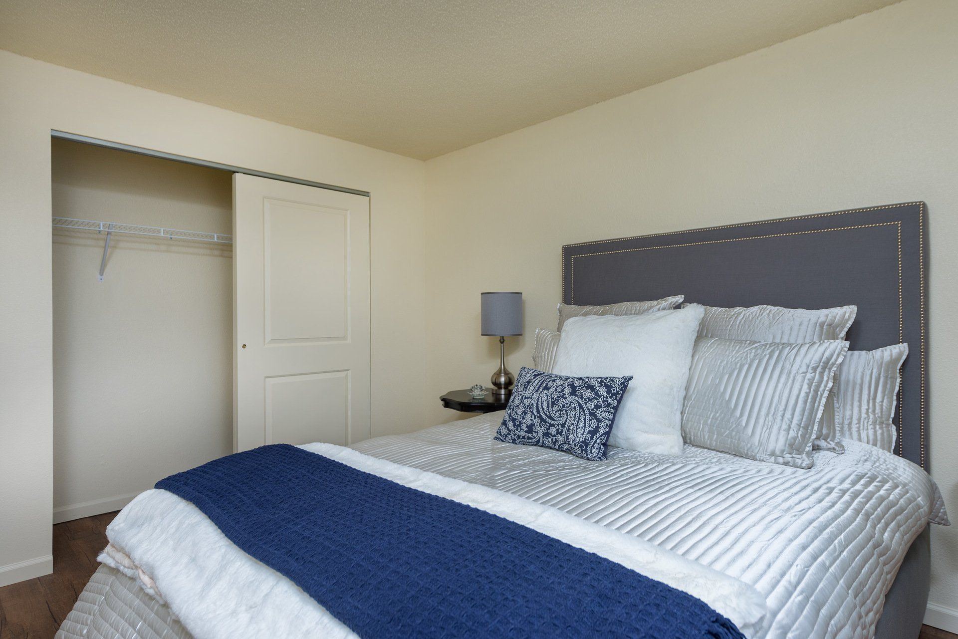 Neat bedroom with gray upholstered bed, blue blanket, nightstand lamp, and open closet door