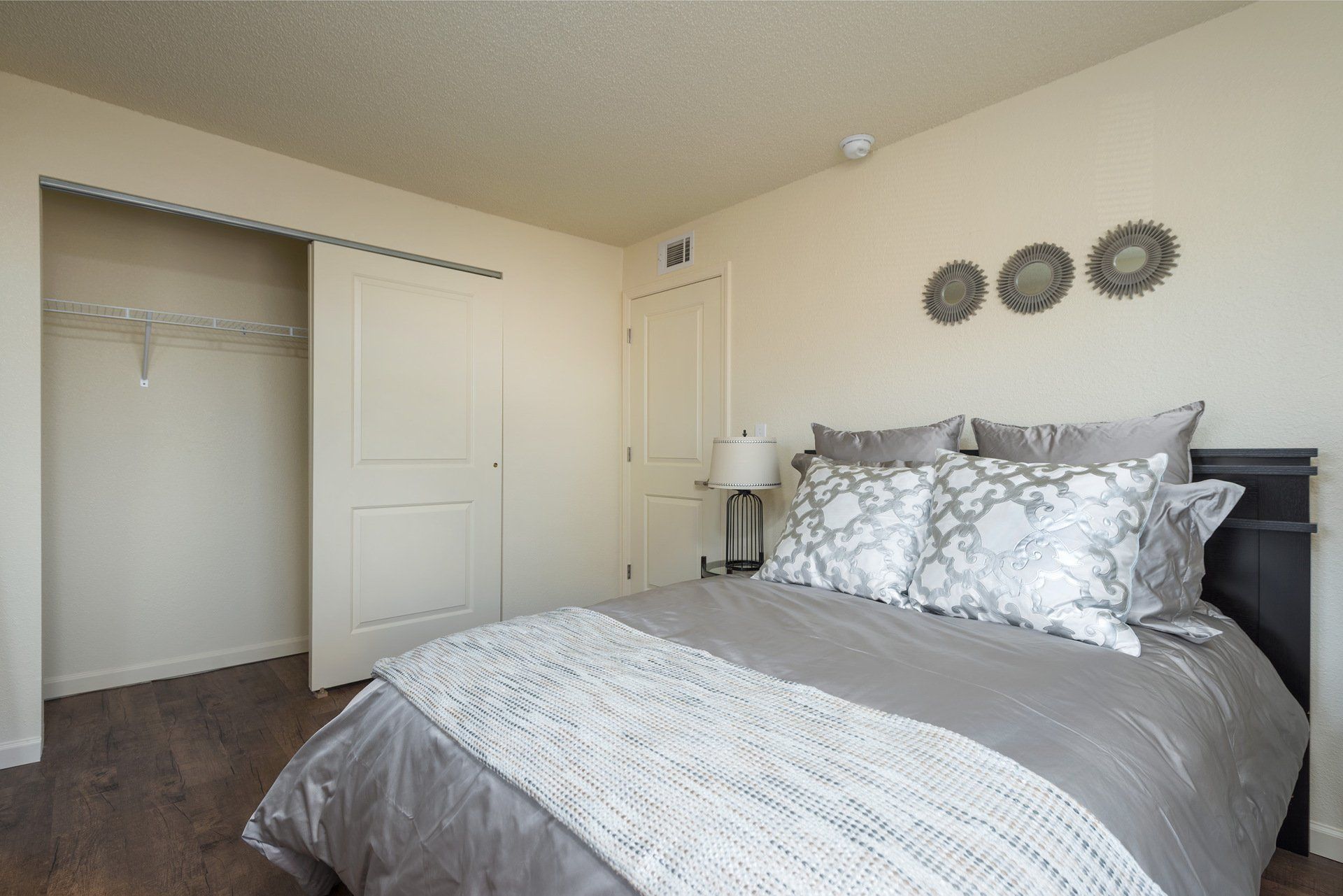 Neatly made bedroom with gray bedding, open closet, and wall decor on a beige wall.