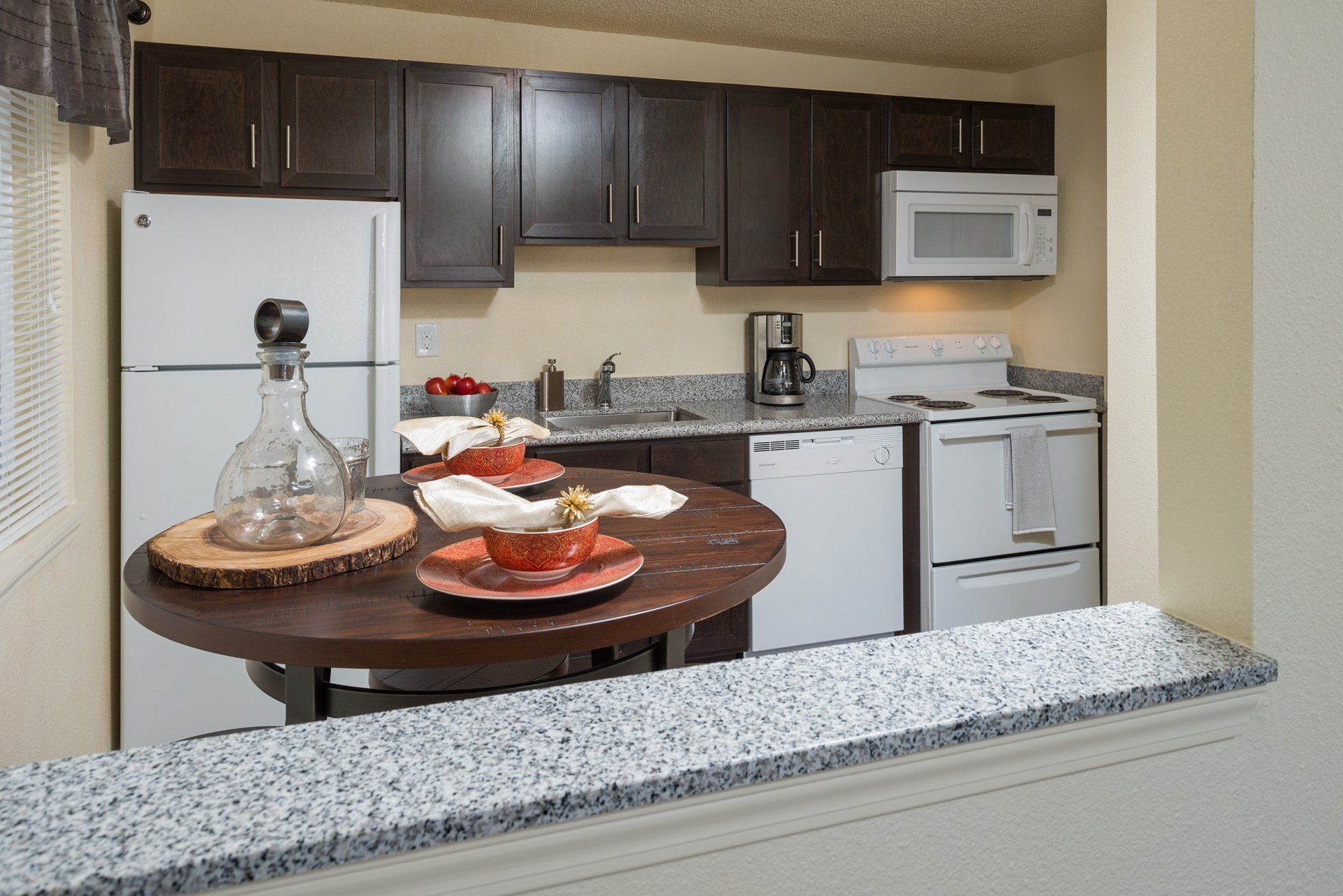 Small kitchen with white appliances, dark cabinets, and a round table set with food and a bottle.