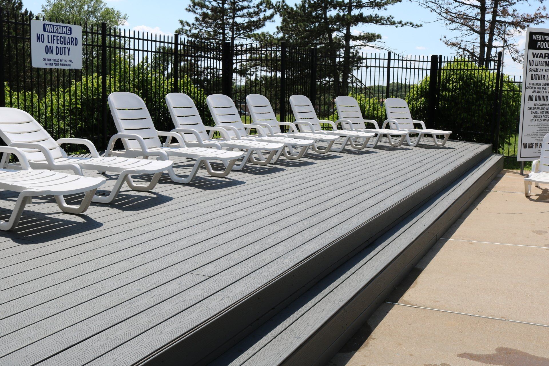 White lounge chairs on a gray pool deck beside a fenced outdoor pool area.