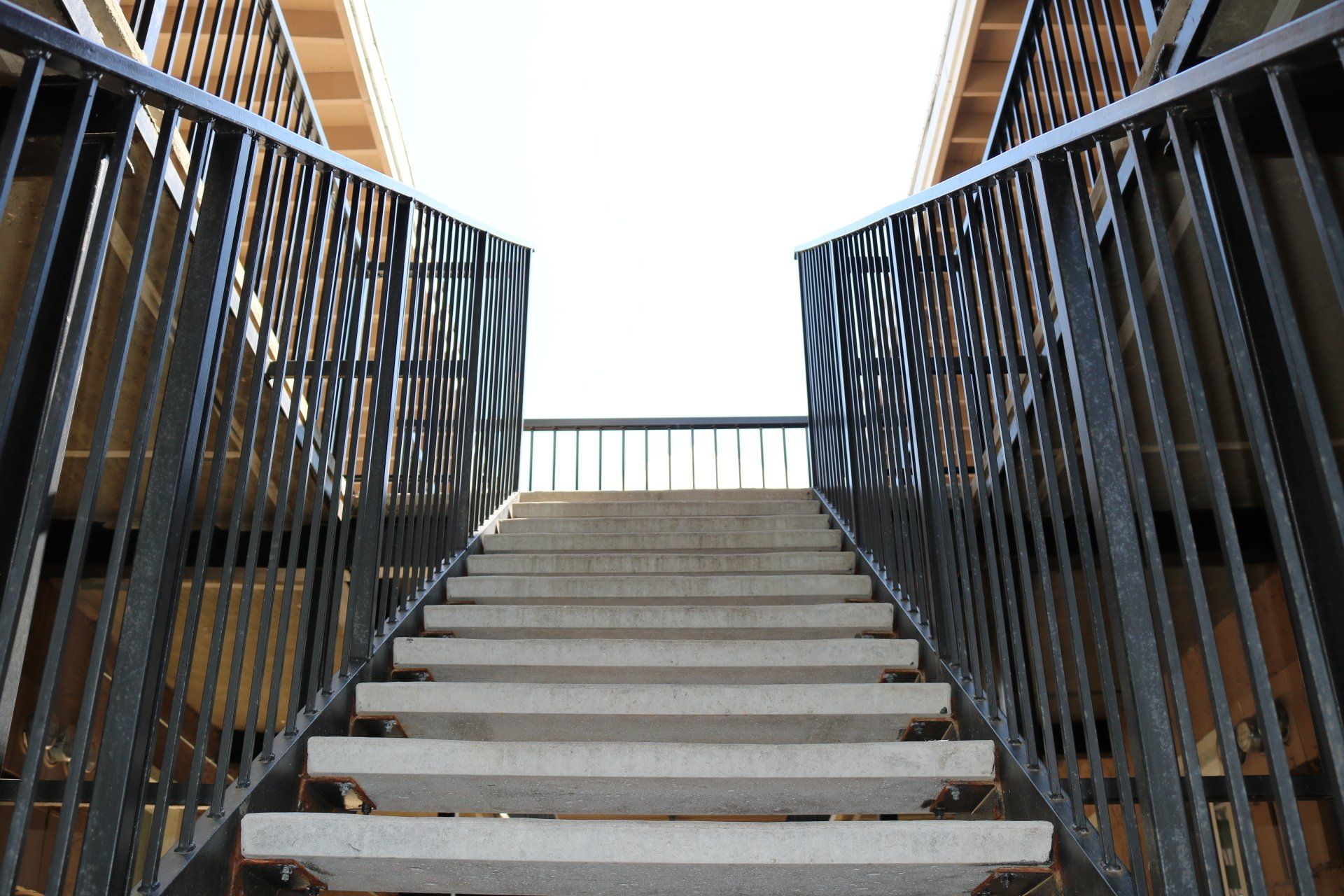 Concrete staircase between black metal railings, viewed from below against a bright sky.