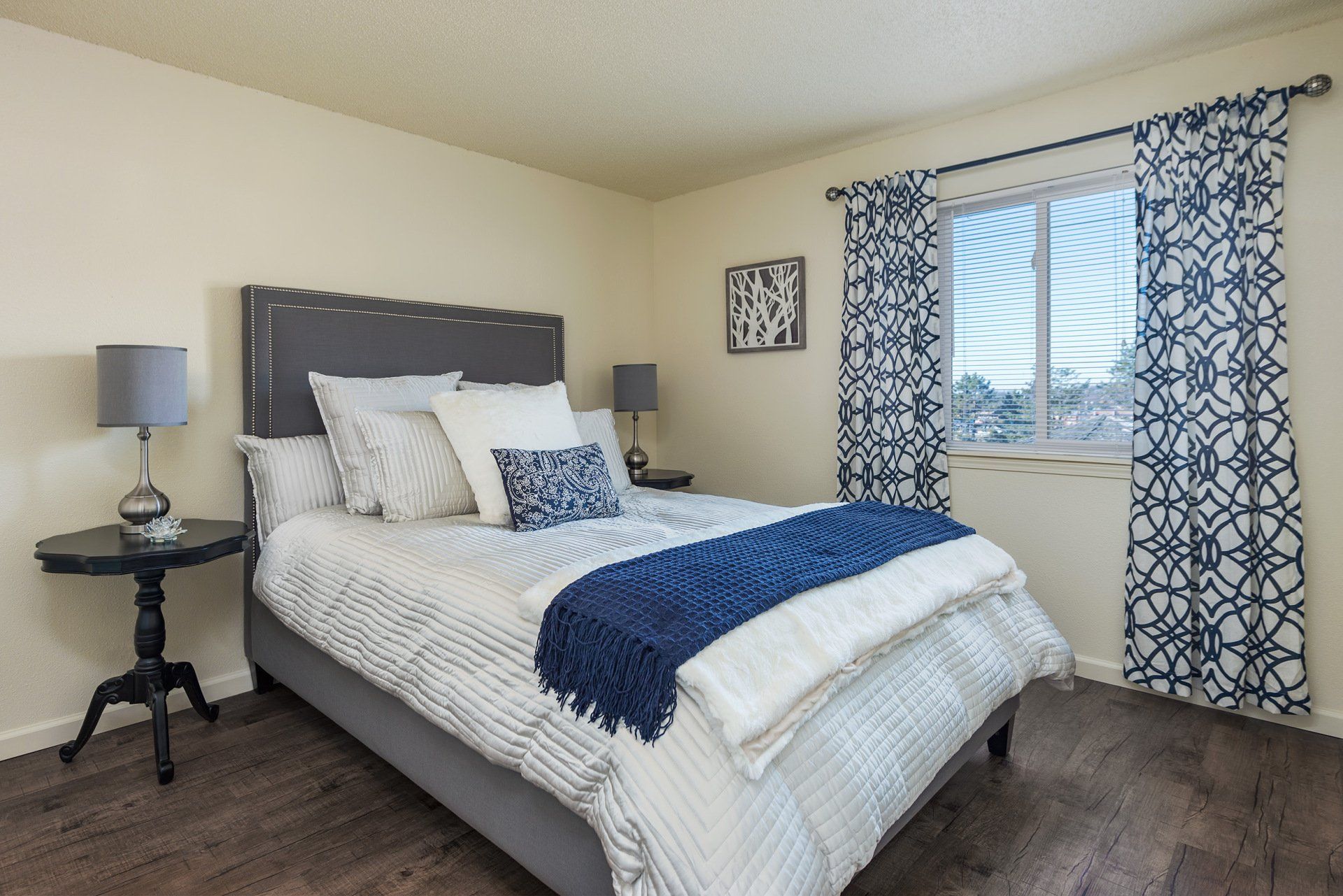 Bright bedroom with a gray bed, blue accents, nightstand lamp, and window with patterned curtains.