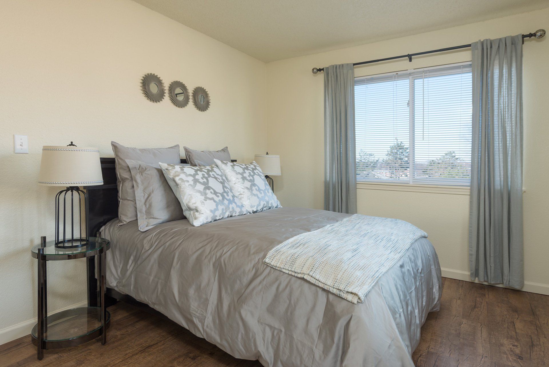 Neatly made bedroom with gray bedding, bedside lamp, wall decor, and a window with curtains.