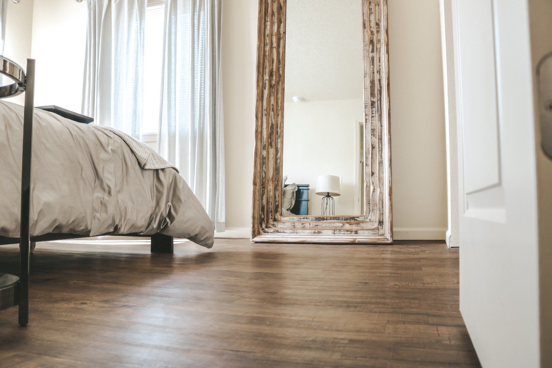 Bright bedroom with wooden floor, bed, and large leaning mirror reflecting a lamp and chair.