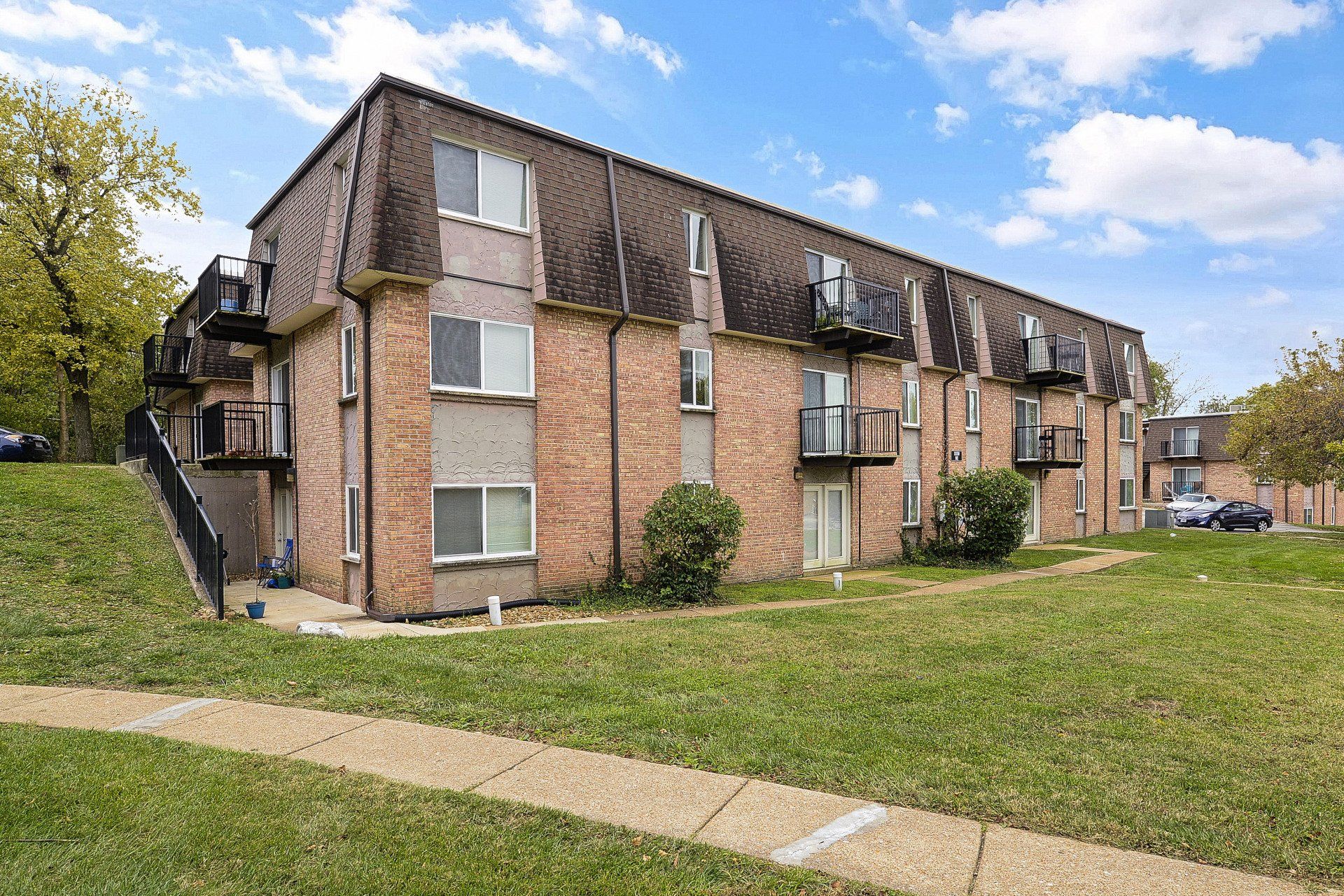 Three-story apartment building with brick and white exterior, grassy lawn, and a sidewalk under a cloudy sky
