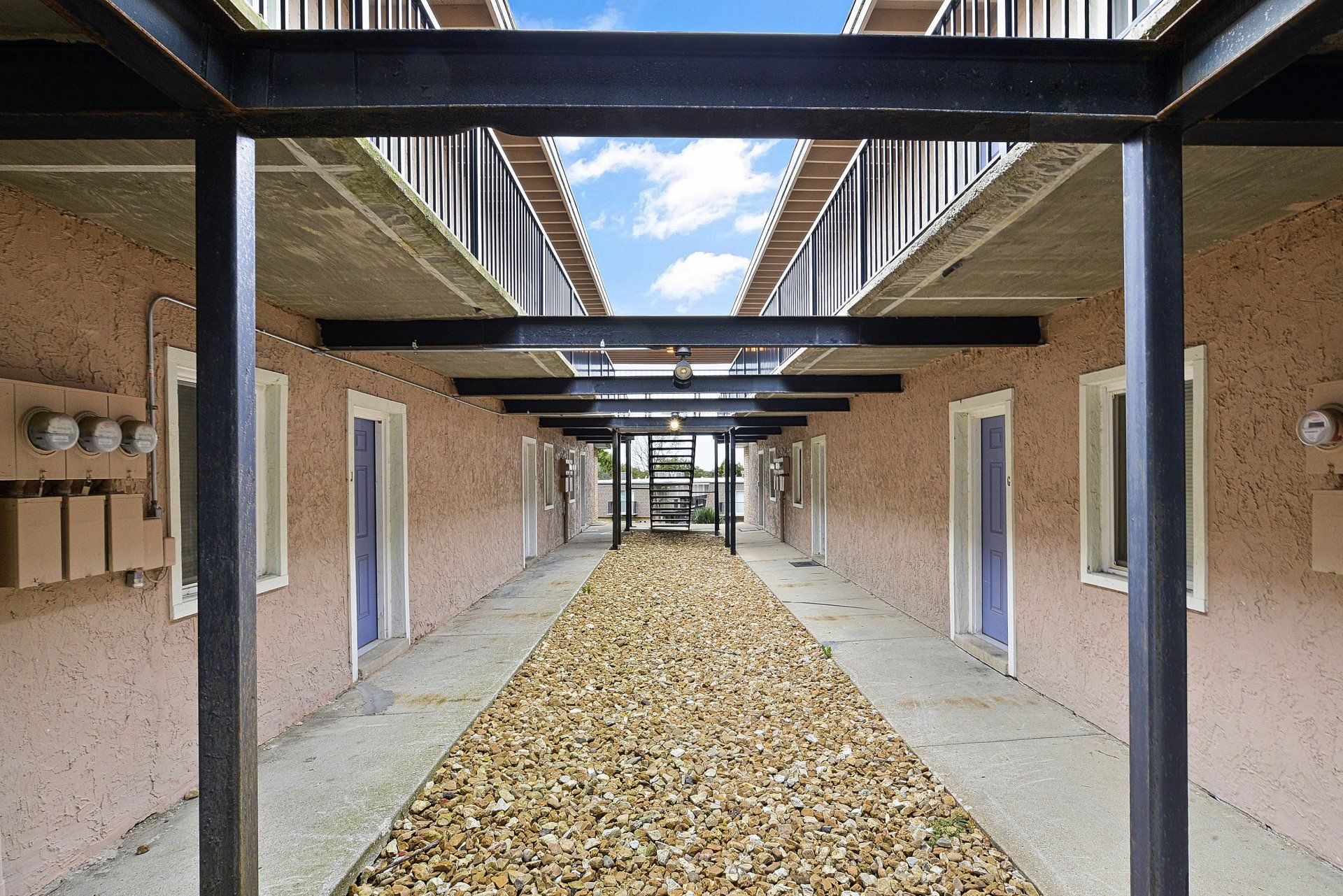 Covered outdoor walkway between tan buildings with black beams and a gravel path