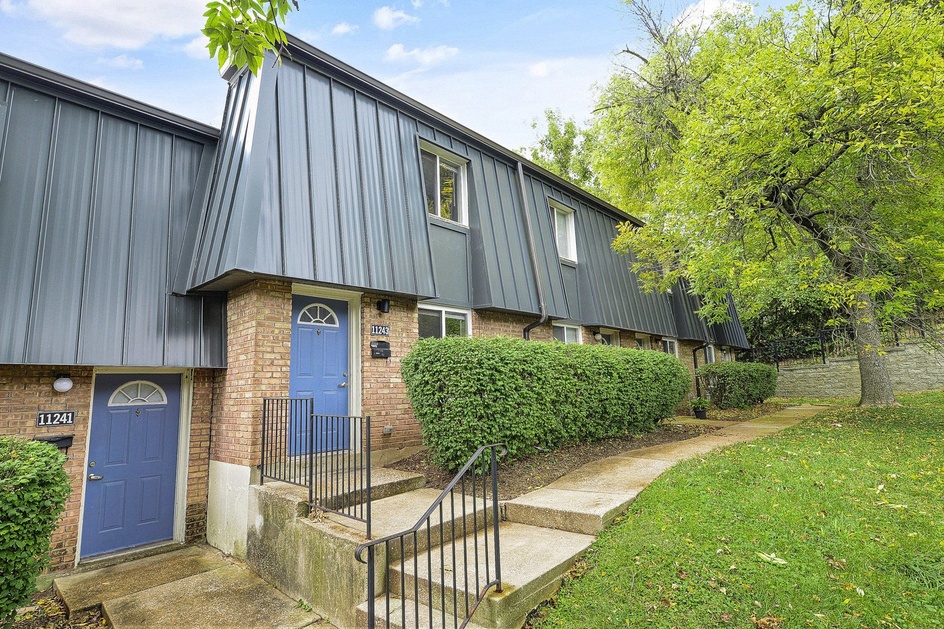 Row of townhouse entrances with blue doors, stone walls, and a sloped grassy path under trees