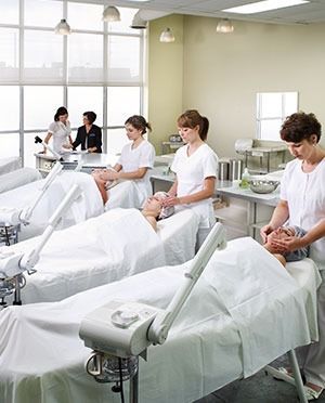 Salon students in white uniforms giving facials in a brightly lit room.
