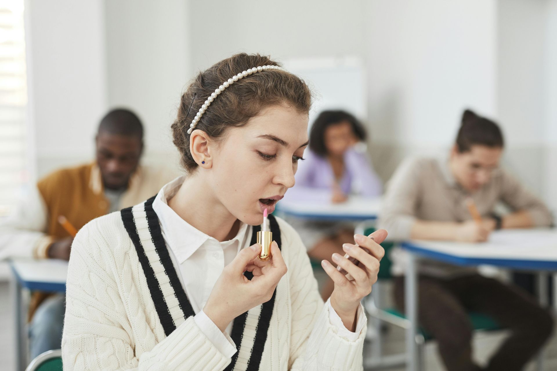 Student applying lipstick at desk during class, with classmates blurred in the background.