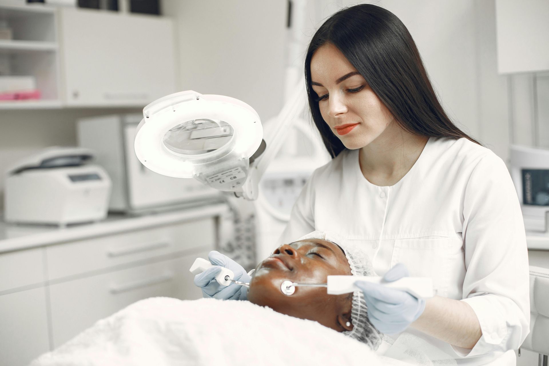 Cosmetologist using a magnifying lamp to treat a client’s face in a bright spa room