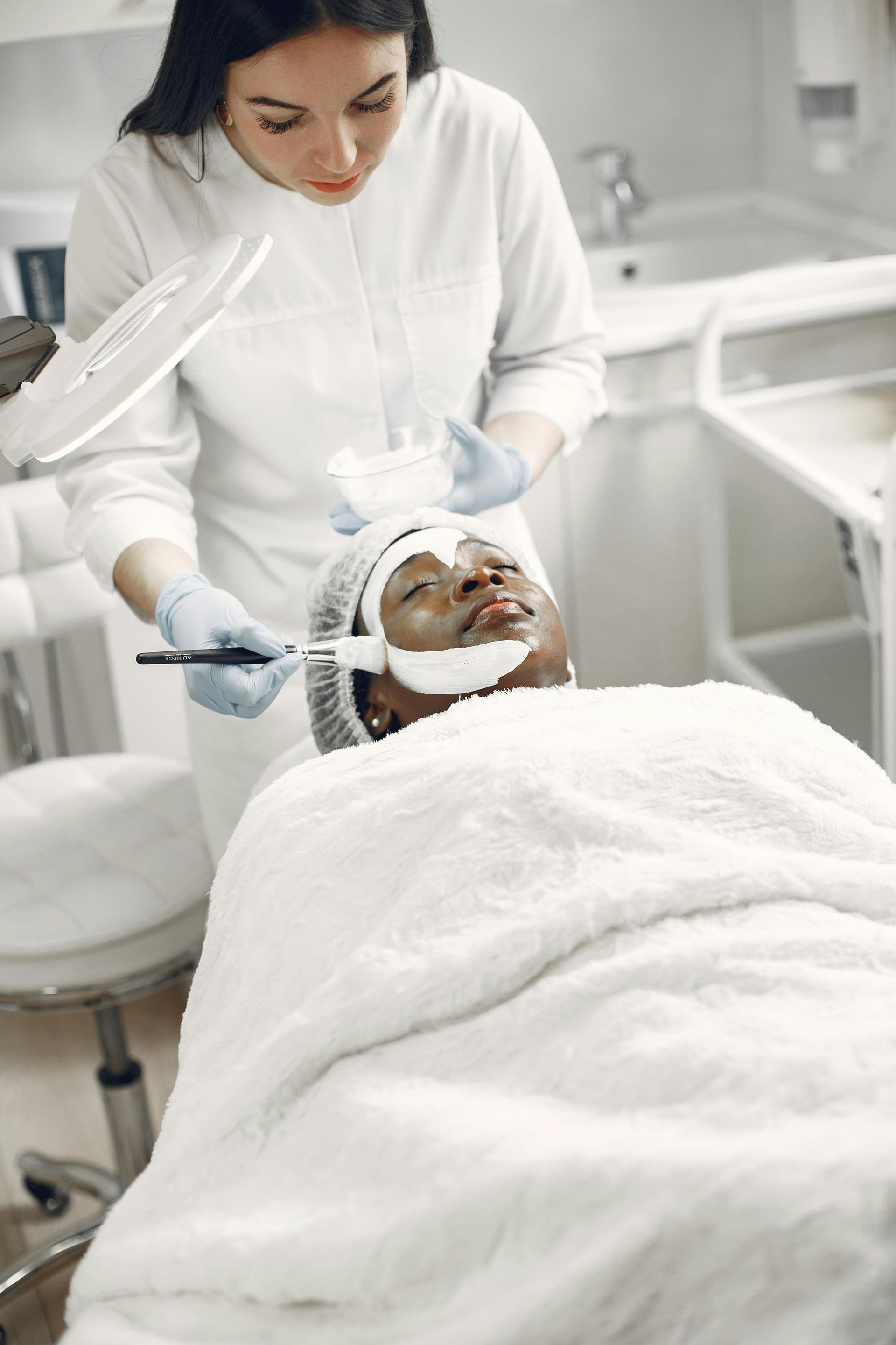 Aesthetician applies facial treatment to a client reclining under a white blanket in a spa room.