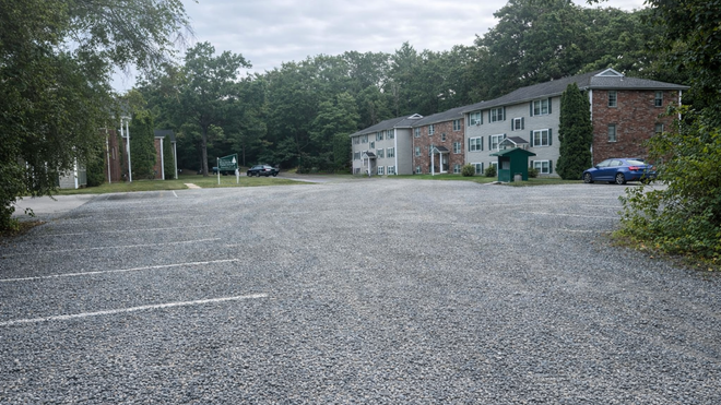 Gravel parking lot in front of a two-story apartment building with a brick tower. Green foliage surrounds the area.