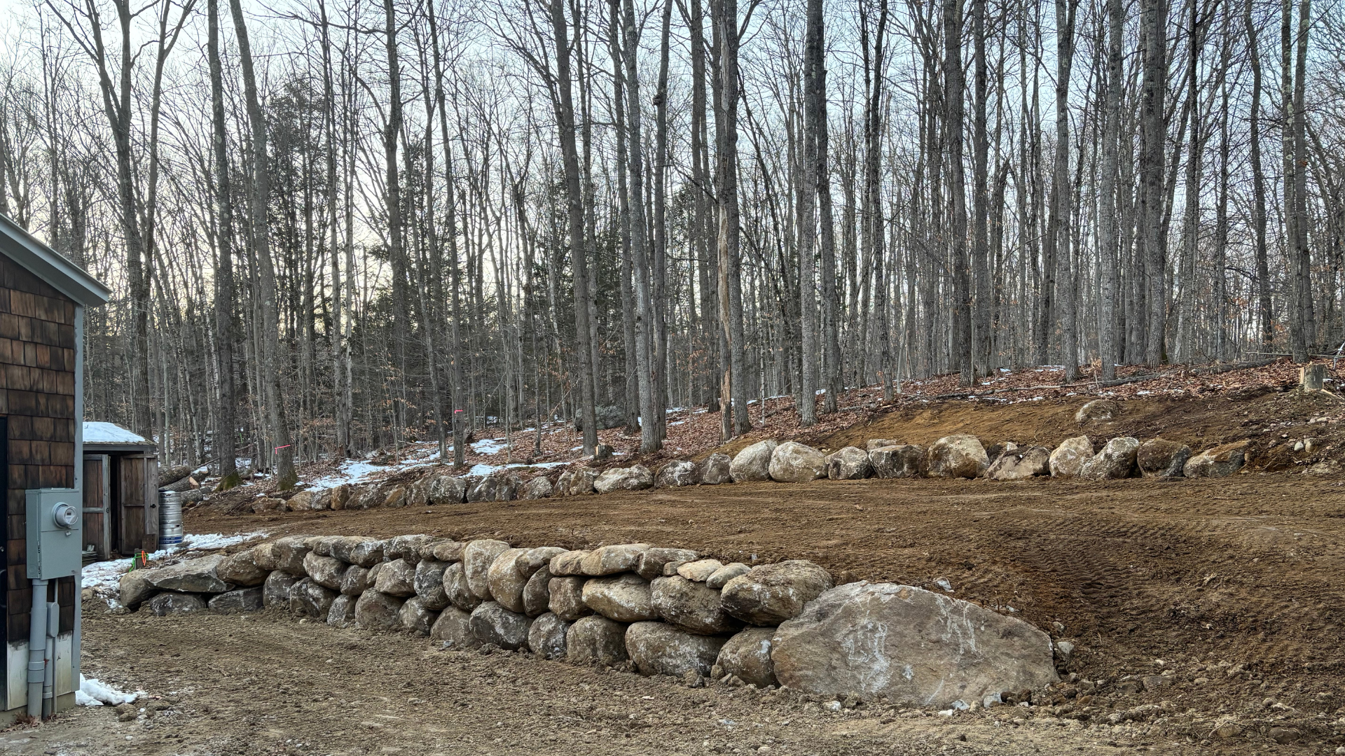 Stone retaining wall on a hillside with bare trees and a building in the background.