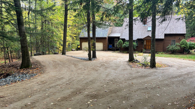 Gravel driveway leading to a log home and detached garage in a wooded setting.