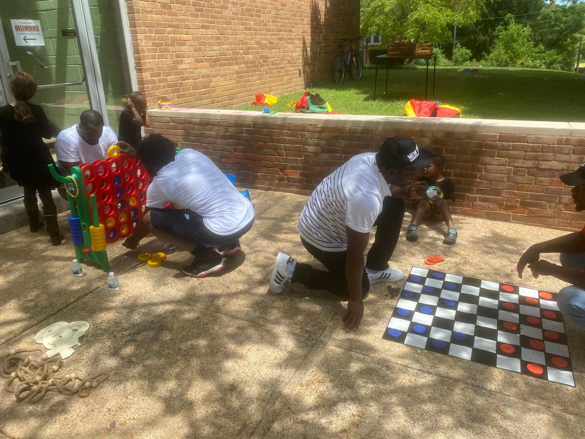 Children kneeling by outdoor checkerboards, playing a game beside a brick wall in sunlight