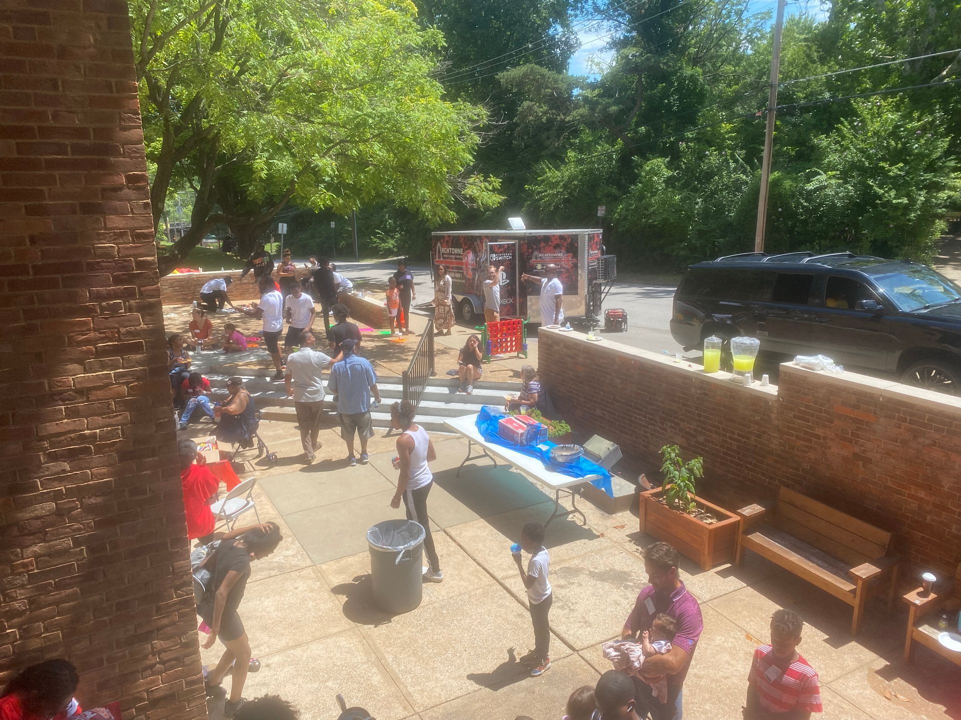 Outdoor courtyard gathering with people seated and standing near tables, a brick wall, and a parked car.
