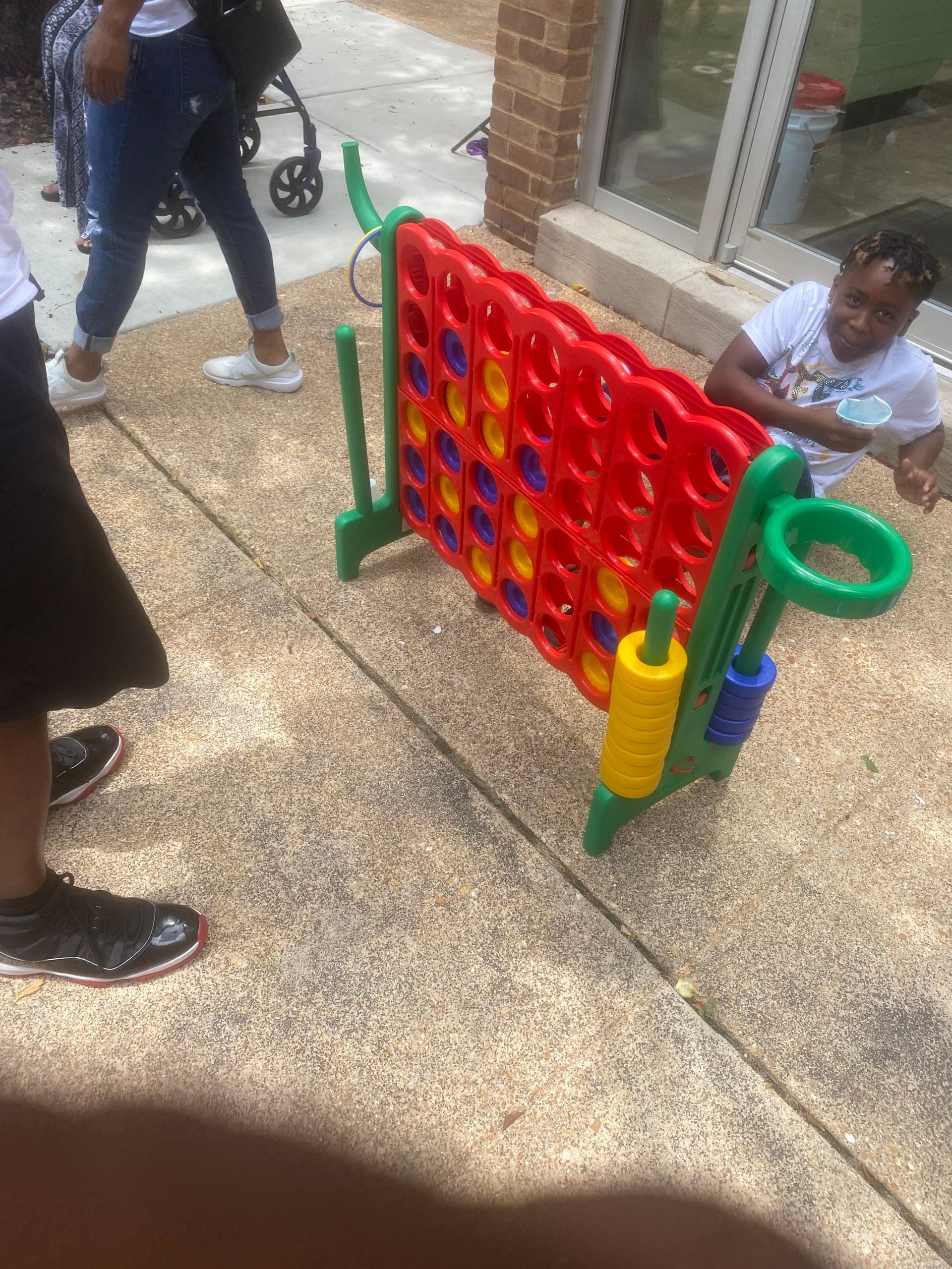 Children playing giant Connect Four on a sidewalk, with a red game board and colorful pieces.