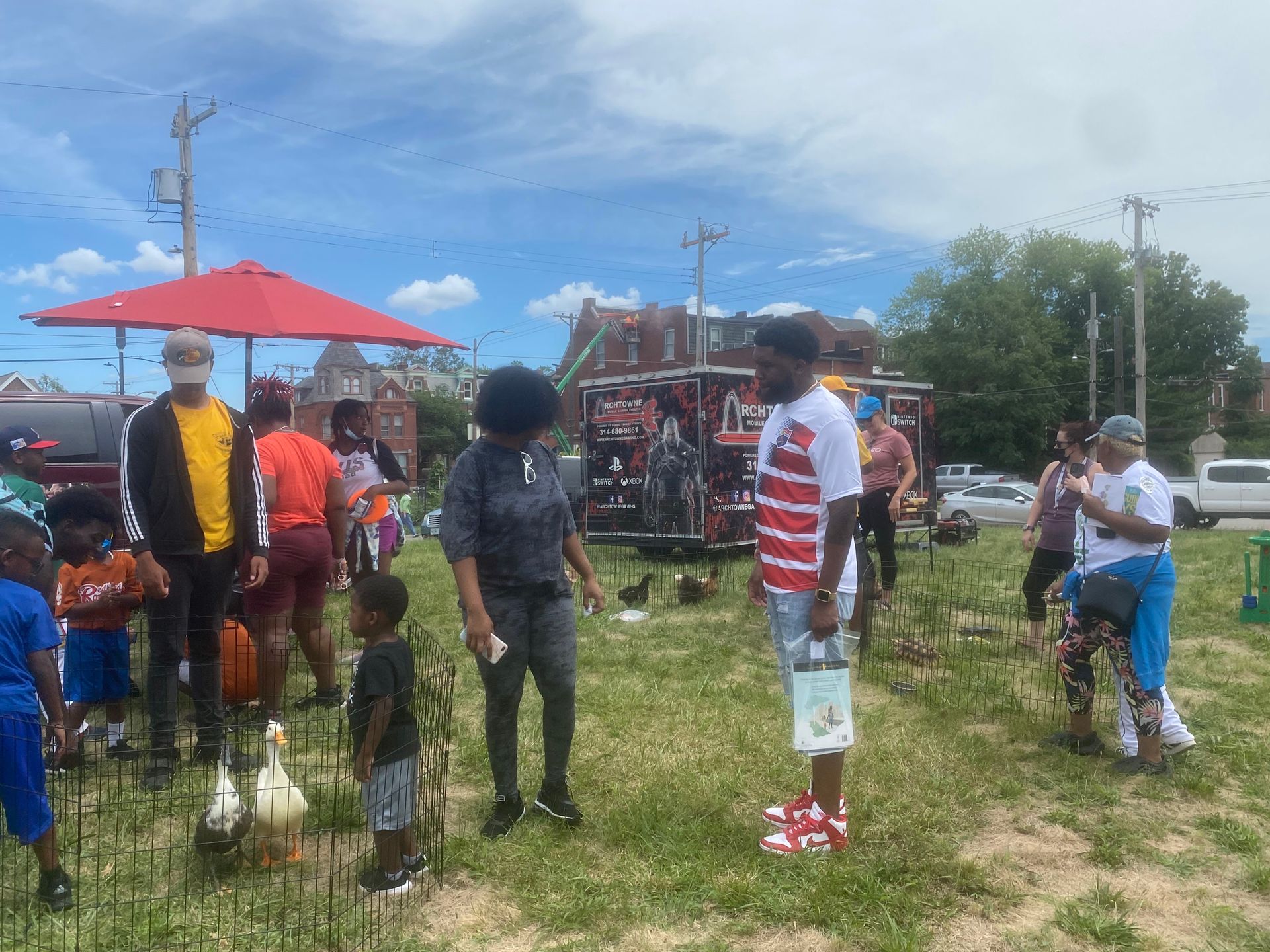 People gathered outdoors near a red umbrella and a red truck at a grassy event.
