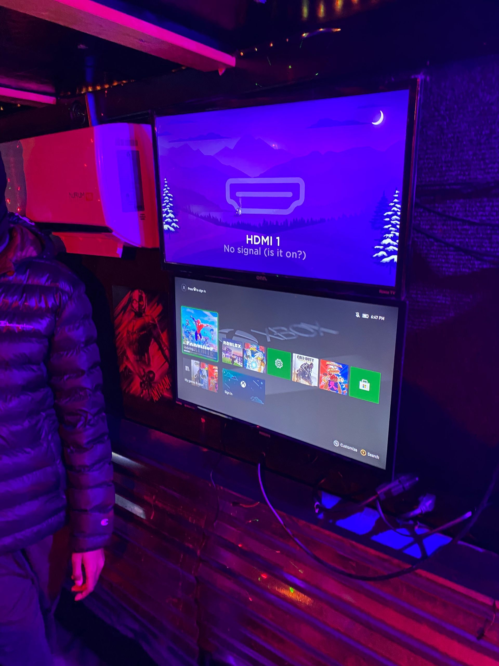 Person beside two monitors in a neon-lit gaming setup with a purple screen and a Windows dashboard.