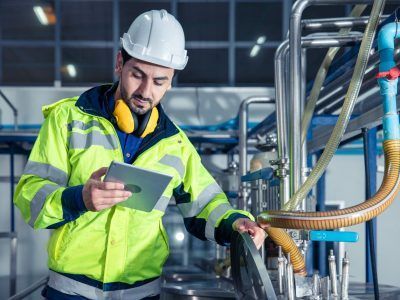 Man in safety gear inspects equipment with a tablet in a factory setting.