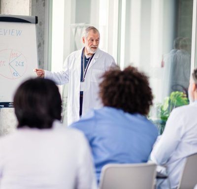 Doctor in lab coat presenting at a seminar, explaining a chart. Other doctors are in the audience.