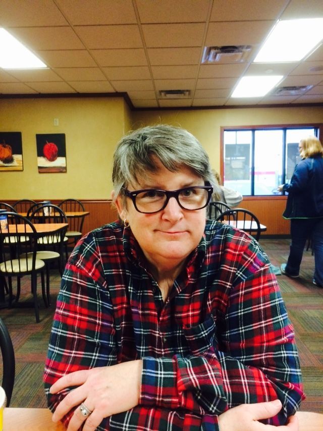 A person with short, gray-brown hair wearing glasses and a red and blue plaid shirt sits at a table in a quiet restaurant.