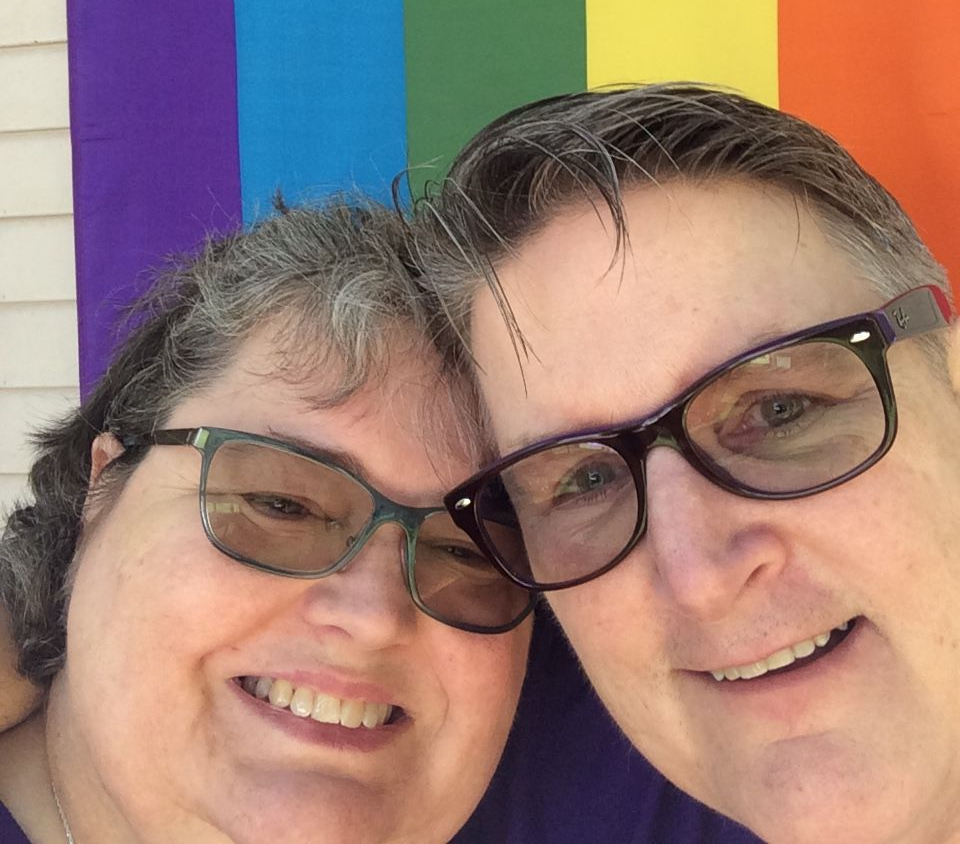 Two smiling people pose head-to-head in front of a pride flag.