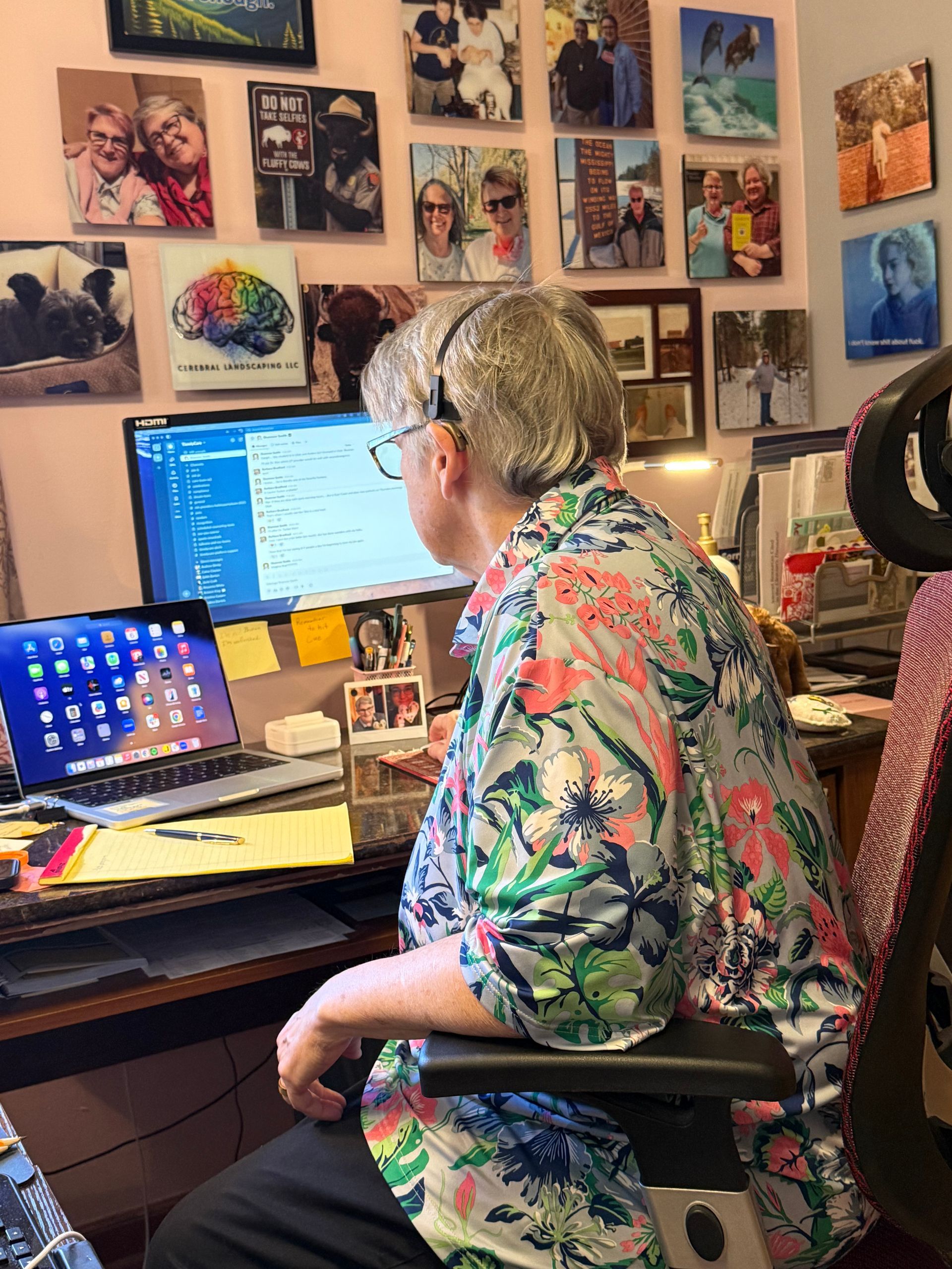 A person with short grey hair wearing a floral shirt sits at a desk with two monitors in a room decorated with many photos.