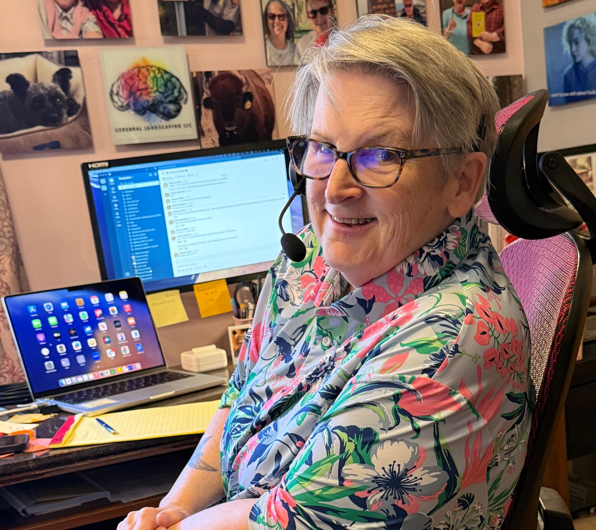 A person wearing a floral shirt and headset sits at a desk with a computer and laptop in a room decorated with wall photos.