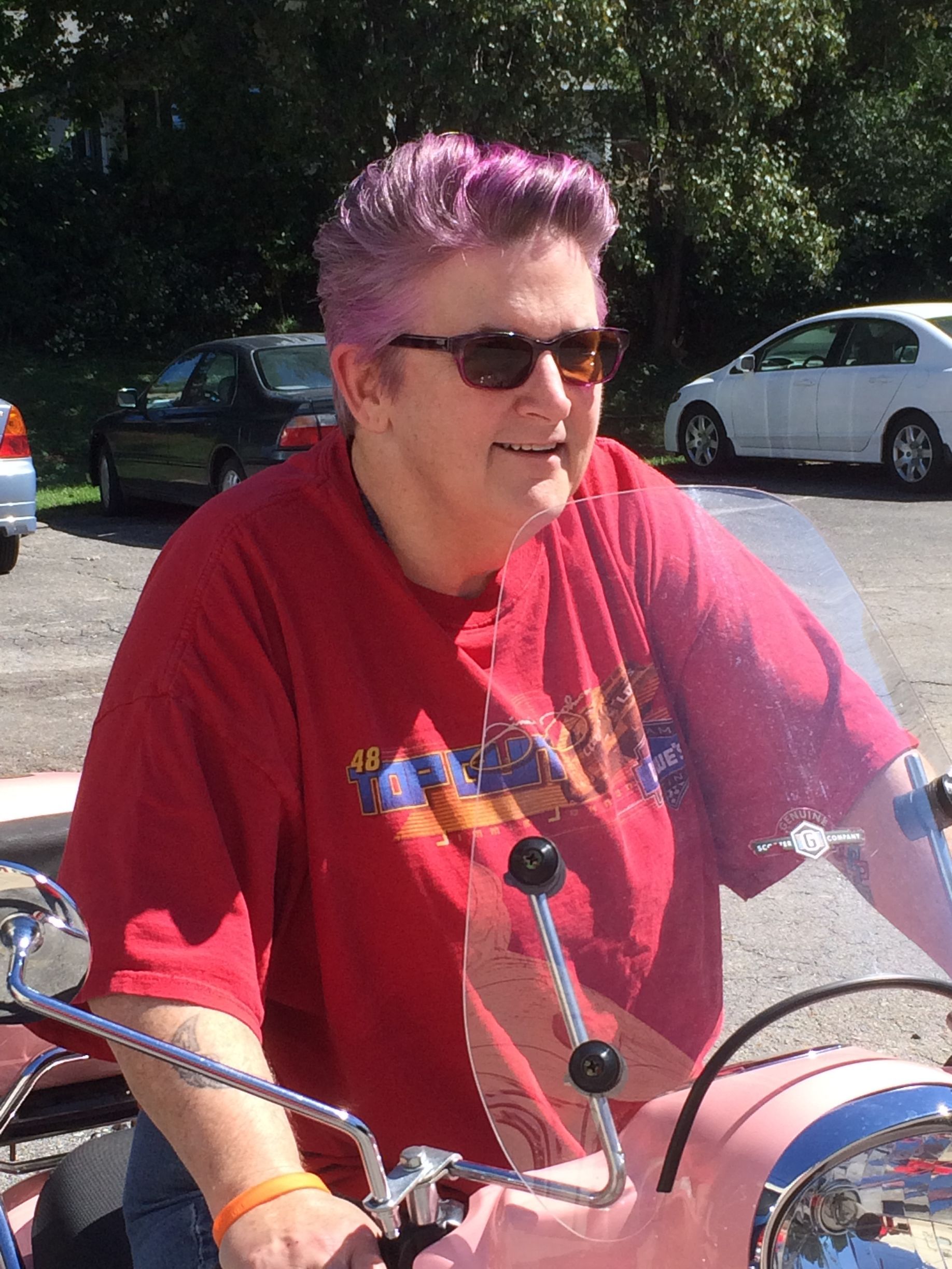 A person with short, magenta-colored hair and sunglasses smiles while sitting on a pink scooter outdoors.