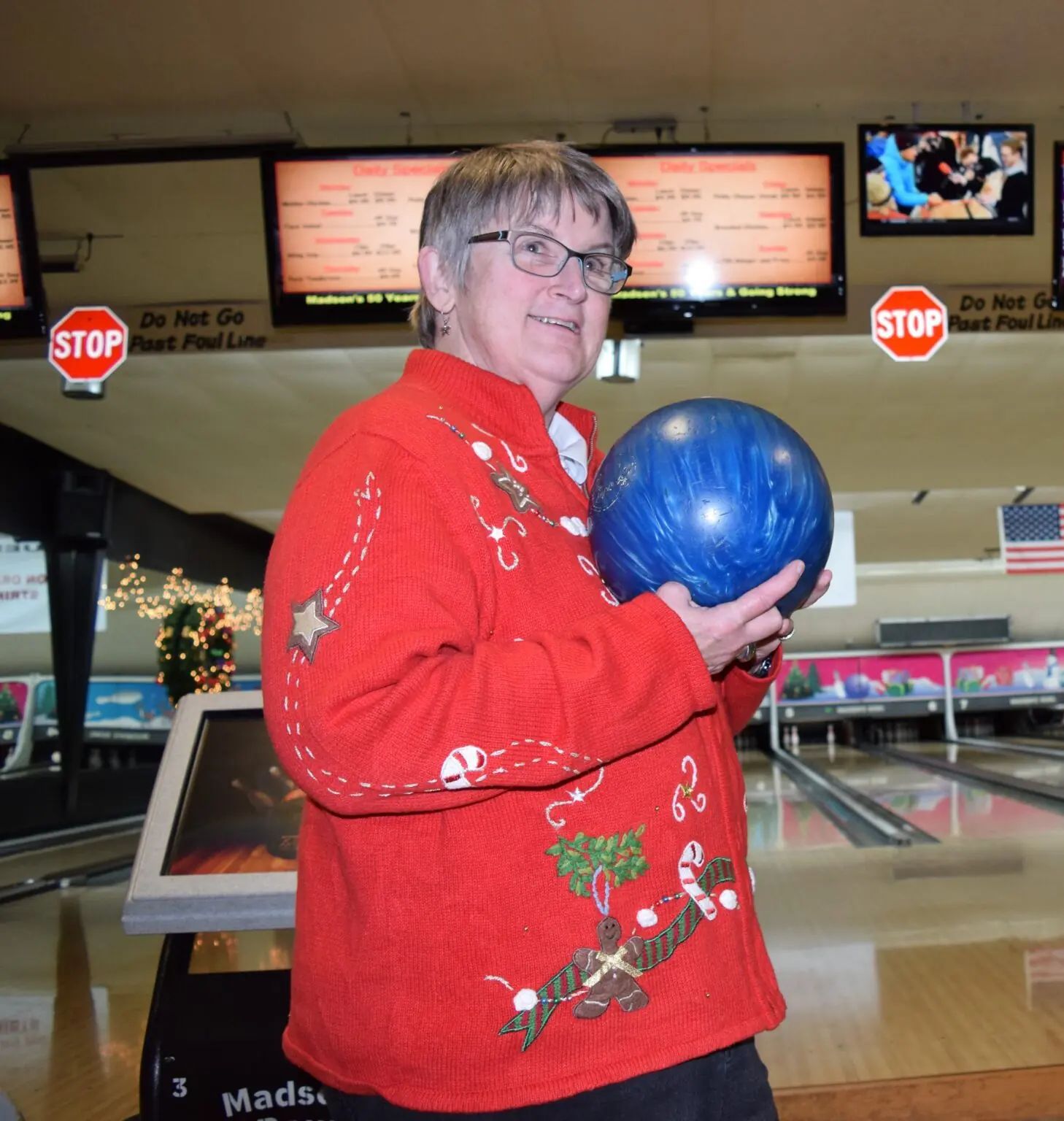 A person in a red Christmas sweater holds a blue bowling ball at a bowling alley with scoreboards in the background.