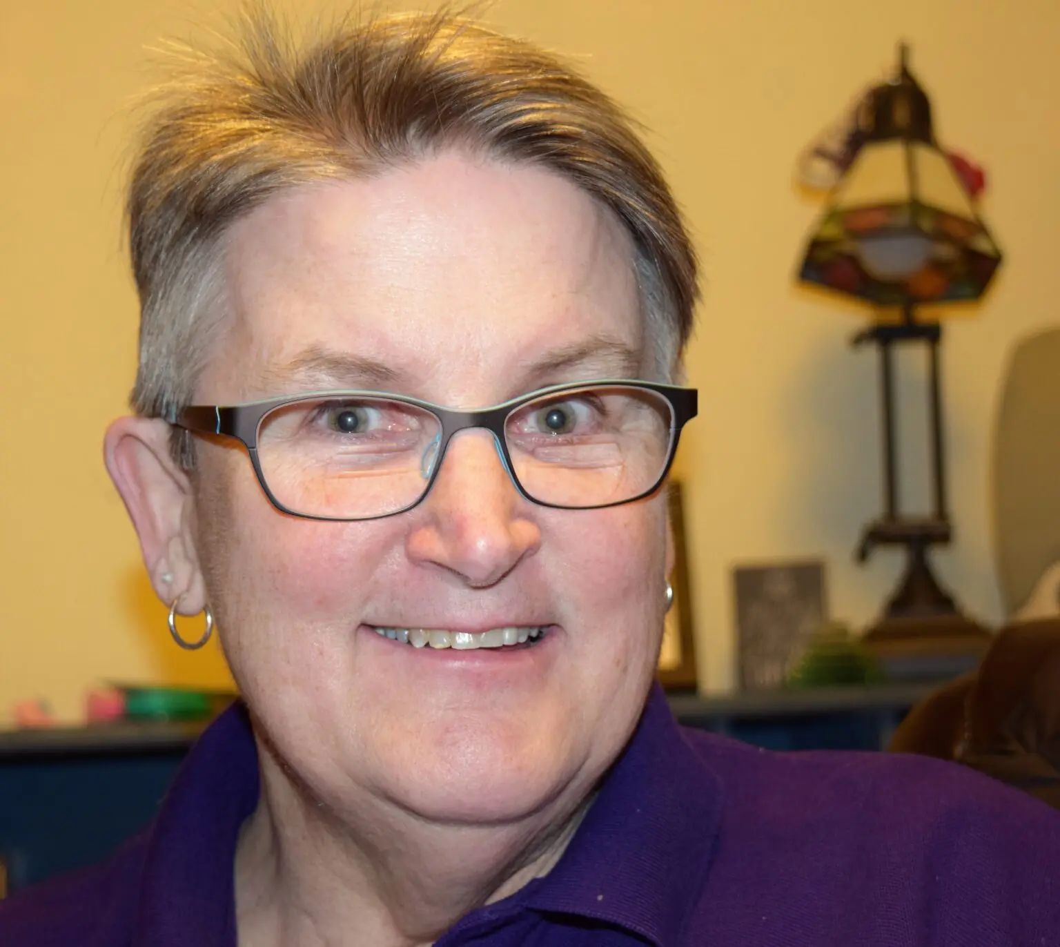 A person with short hair and glasses wearing a purple collared shirt, smiling in an indoor setting.