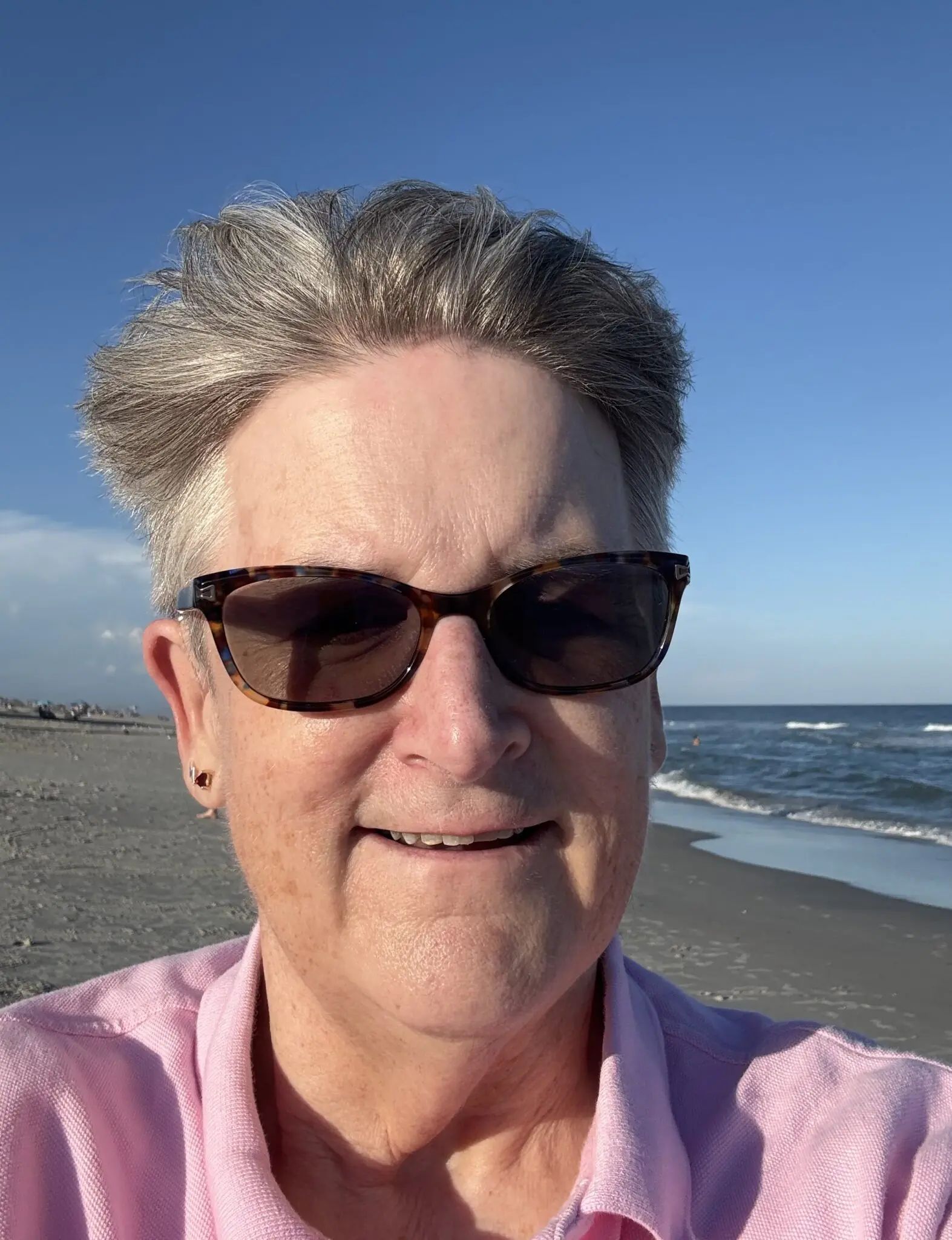 A person wearing sunglasses and a pink collared shirt smiling on a beach with the ocean in the background.