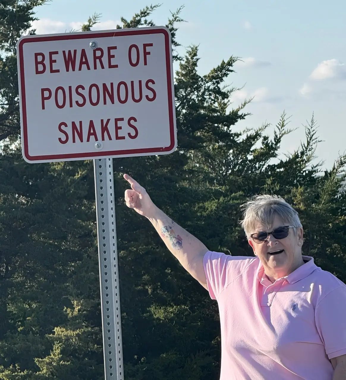 A person in a pink shirt smiling and pointing to a sign that says, 