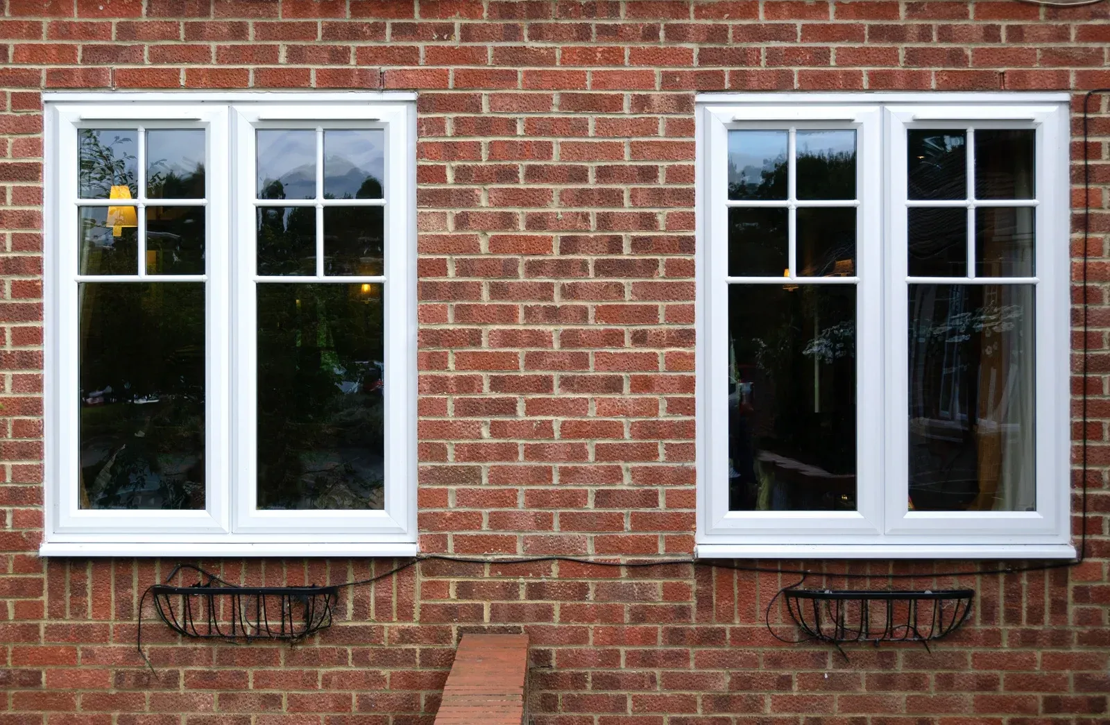 Two white-framed windows on a brick wall, each with a small black planter below.