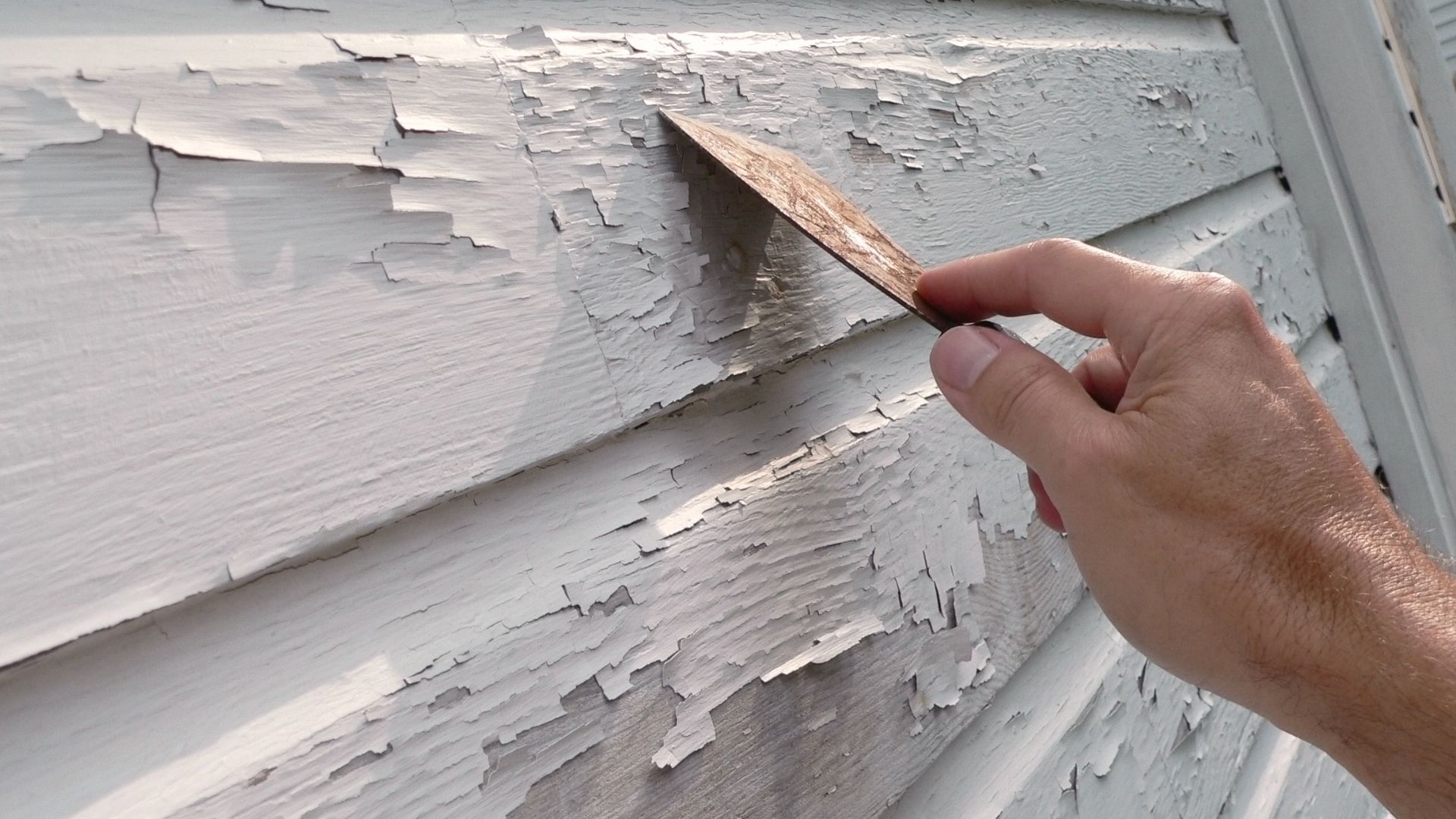 Hand scraping peeling white paint from weathered wood siding with a putty knife